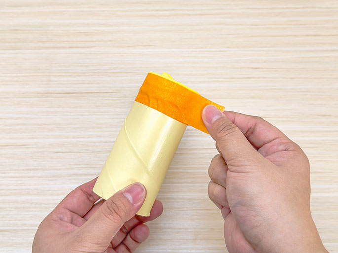 Two hands hold a pale yellow cardboard tube over a light wooden surface. One hand holds the tube while the other applies a piece of orange masking tape around the top edge. The process of taping the tube is the central focus of the image.