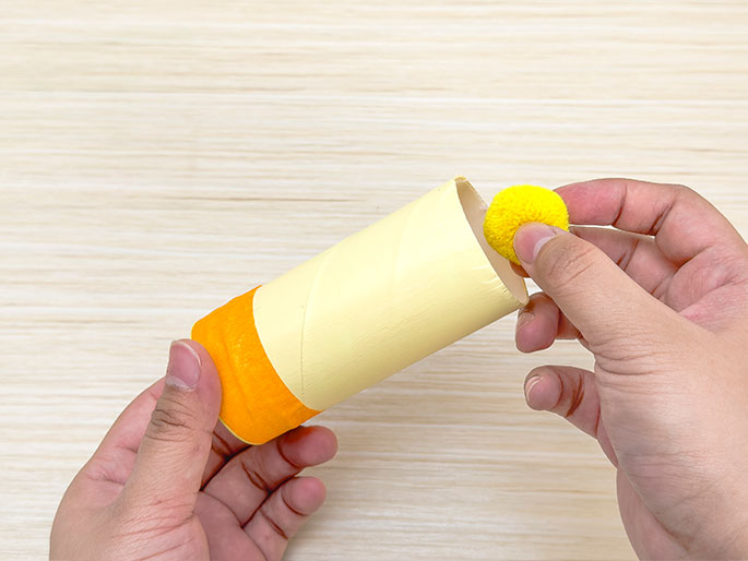 Two hands manipulate a cardboard tube painted pale yellow with an orange band of tape around one end. One hand holds the tube, and the other hand places a bright yellow pom-pom into the open end of the tube, against a light wooden background. The image suggests a crafting activity.