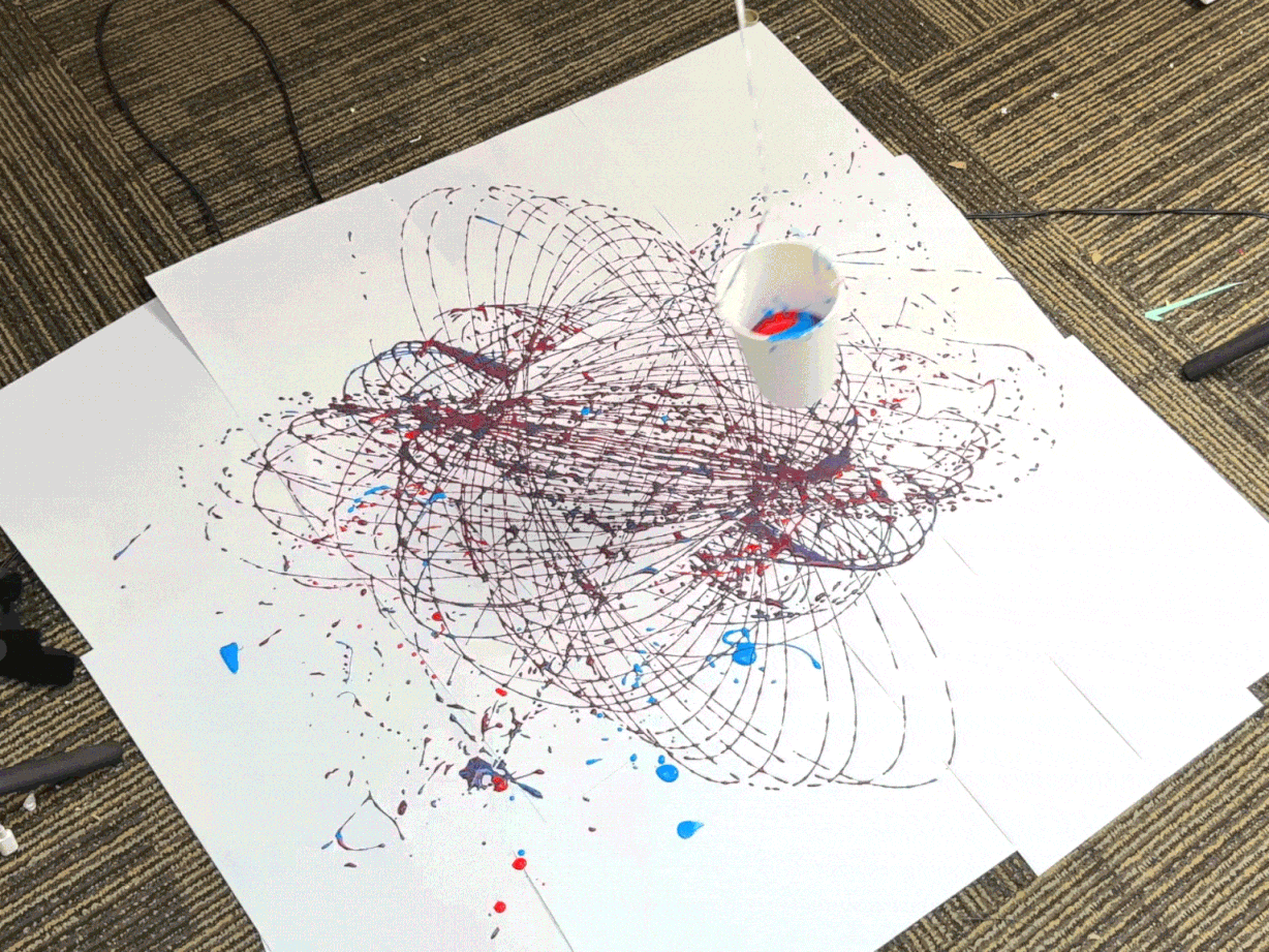 A small cup or container, likely punctured with holes, hangs suspended above a large piece of white paper. The cup contains red and blue paint, which drips and splatters onto the paper below. The cup swings in a pendulum motion, creating swirling and overlapping patterns of red and blue lines and splatters on the paper. The video captures the process of creating abstract art using a swinging pendulum and paint.