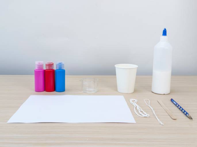 An assortment of craft supplies is arranged on a light wood-grained table against a plain off-white background. Three small bottles of brightly colored paint—pink, red, and blue—are grouped together. Next to them is a small, clear glass or plastic measuring cup. A white paper cup, a squeeze bottle of white glue, a piece of white paper, two lengths of white string, a small wooden craft stick, and a pencil complete the collection of materials. The scene suggests preparation for a children's art or craft project.