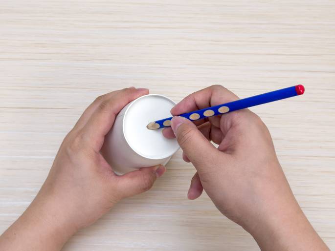 Two hands interact with a white paper cup on a light wood-grained surface. One hand holds the cup upside down, presenting its bottom. The other hand holds a blue pencil with a red eraser and uses it to poke or create a hole in the center of the cup's base. The focus is on the action of creating the hole, suggesting a craft or DIY activity.