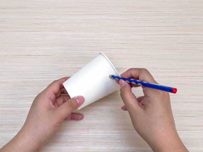 On a light wood-grained surface, two hands are shown interacting with a plain white paper cup. One hand holds the cup on its side, while the other hand, holding a blue pencil with a red eraser and dotted grip marks, makes a mark or small hole on the side of the cup. This suggests the cup is being prepared for a craft project or some other modification.