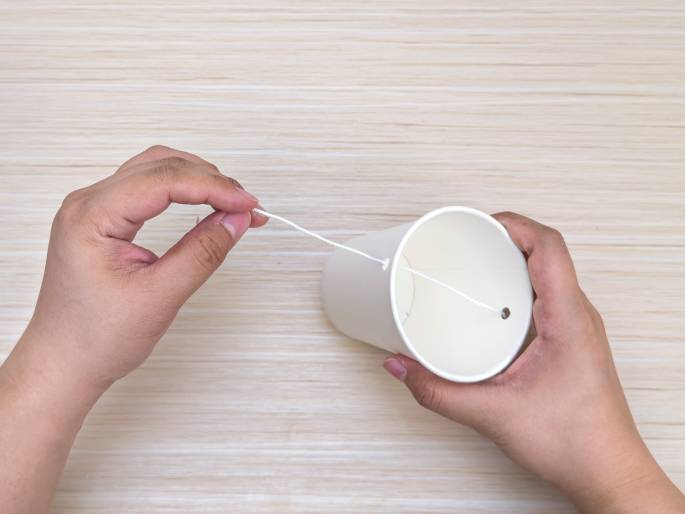 Two hands are working with a white paper cup and a piece of white string on a light wood-grained table. One hand holds the cup, which has a small hole near the bottom edge. A piece of string is threaded through this hole and extends out. The other hand gently pinches the string. The image appears to depict the process of creating a simple toy or craft, possibly a string telephone or a small hanging decoration.