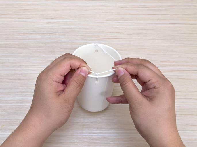 Two hands manipulate a white string attached to a white paper cup on a light wood-grained table. The cup has two small holes near the top edge, and the string is threaded through these holes, creating a loop or handle. The hands are adjusting the string, likely tightening or securing the loop. This suggests the construction of a simple hanging container or a component of a craft project.