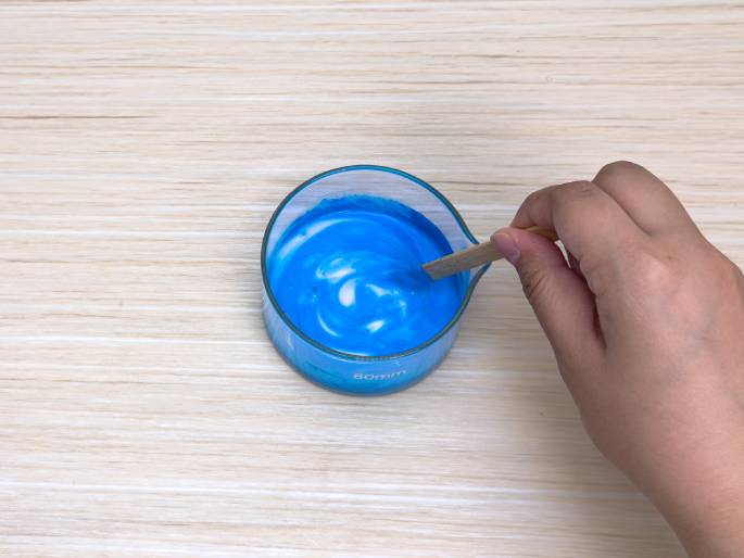 A hand uses a small wooden stick to stir a vibrant blue substance within a clear glass container on a light wood-grained surface. The container appears to be a measuring beaker or cup, and the blue substance has a slightly viscous or thick consistency. The image suggests the mixing of paint, dye, or some type of craft material.