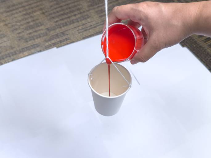 Against a white background, a hand pours a thick, red liquid from a clear container into a small white paper cup suspended by strings. The red liquid streams down into the cup. The setup resembles a miniature bucket being filled, possibly for a science experiment or craft activity. A textured brown surface is visible at the top edge of the image.