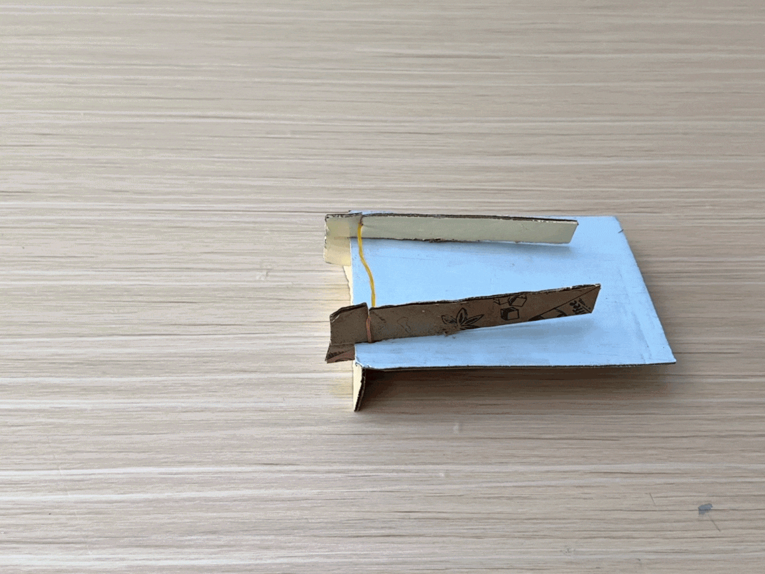 Hands operate a small catapult constructed from cardboard and a rubber band. A red cardboard projectile is launched from the device across a wooden surface.