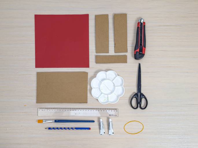 An overhead, flat lay view displays various craft supplies arranged on a light wood-grain surface. The supplies include: a sheet of red paper or cardstock, pieces of brown cardboard, a utility knife with a red handle, a pair of black scissors, a white flower-shaped paint palette, a clear plastic ruler, a blue paintbrush, a blue pencil, two small tubes of white glue or paint, and a yellow rubber band. The arrangement suggests preparation for a craft or art project.