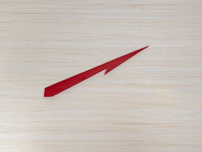 A small, angular piece of red paper, resembling a stylized arrowhead or a shard, lies on a light wood-grain background. It appears to be a scrap or offcut, possibly from a previous crafting step.