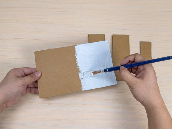 Two hands are painting a piece of cardboard white with a paintbrush on a light wood-grain background. One hand holds the rectangular piece of cardboard while the other hand applies the white paint, covering approximately half of the cardboard's surface. Three additional, smaller pieces of unpainted cardboard are visible to the upper right, suggesting they are also part of the project. This image likely depicts a stage in a crafting or DIY project.