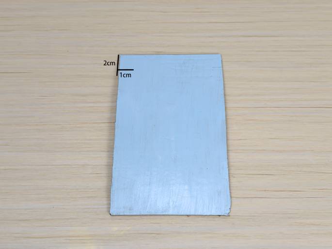 A rectangular piece of light blue painted cardboard sits on a light wood-grain background. Lines and labels indicate measurements: 2cm along the shorter side and 1cm along the longer side, suggesting a small piece of cardboard. The paint appears slightly uneven, indicating a hand-painted finish. This image likely shows a prepared piece for a craft or DIY project.