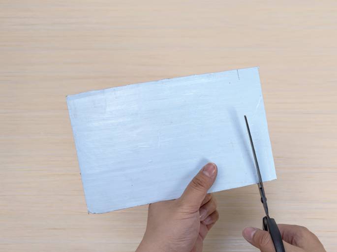 Two hands are shown cutting a piece of light blue painted cardboard with scissors on a light wood-grain background. One hand holds the rectangular cardboard piece while the other hand operates the scissors, beginning to make a cut along one of the shorter edges. This image likely depicts a step in a crafting or DIY process.