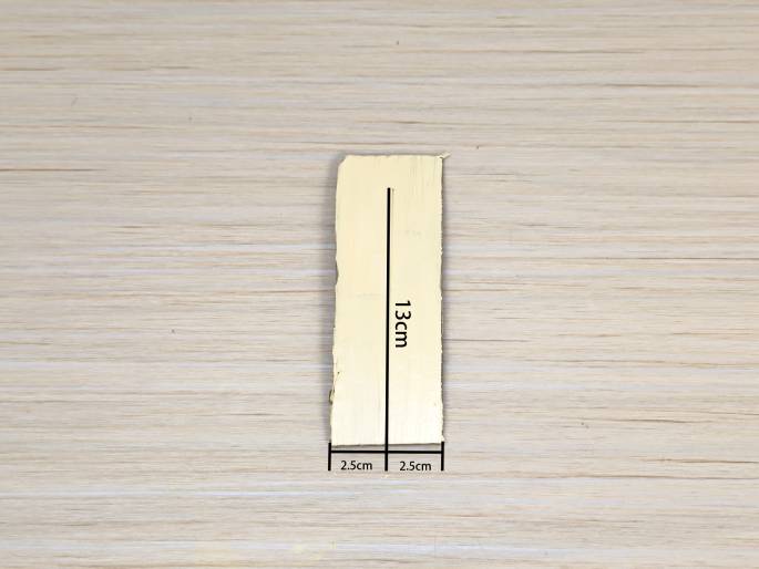 A long, thin strip of light-colored cardboard or wood lies on a light wood-grain background. Lines and labels indicate its dimensions: 13cm along the length and 2.5cm along the width at both ends. This image likely shows a piece prepared for a craft or construction project.