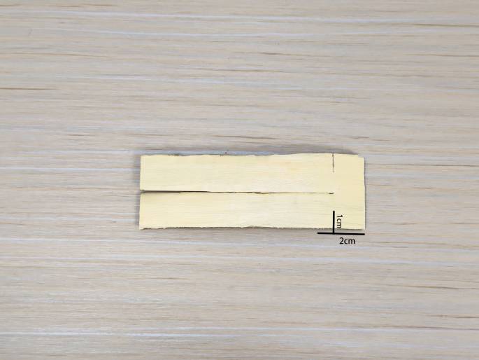A small, light-colored piece of cardboard or wood, partially cut, is placed on a light wood-grain background. The piece has a narrow slit cut almost all the way through, dividing it into two thin sections. Lines and labels indicate measurements: 2cm along the bottom edge and 1cm for the width of the uncut portion. This image likely illustrates a step in a craft or model-making project.