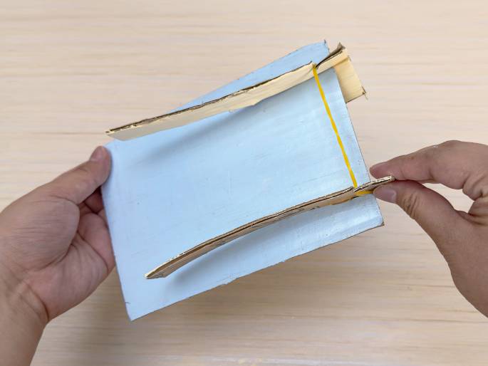 Two hands are assembling a small structure made of light blue painted cardboard and thin, unpainted cardboard strips against a light wood-grain background. A yellow rubber band is stretched between the two upright cardboard strips, which are attached to the blue base. One hand holds the base while the other hand adjusts one of the cardboard strips, suggesting the process of building a small catapult or similar device.