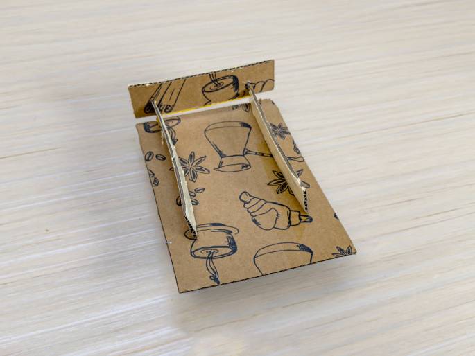 A small catapult or similar device, constructed from cardboard, is displayed on a light wood-grain background. The base of the device is a piece of cardboard decorated with a repeating pattern of coffee-related images, including coffee beans, croissants, cinnamon sticks, and coffee pots. Two thin, unpainted cardboard strips are attached to the base and angled upwards, connected at the top by a small strip of the same cardboard. This image likely showcases a finished DIY project, possibly a toy or a decorative item.