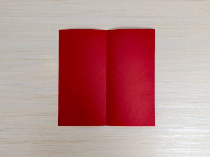 A rectangular piece of red paper or cardstock is folded in half, creating a vertical crease down the middle. It lies open, book-style, against a light wood-grain background. The image likely depicts a step in an origami or paper craft project.