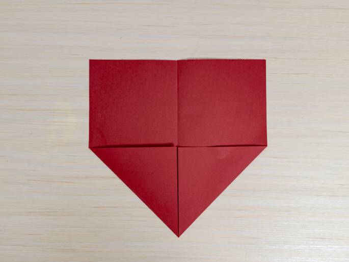 A piece of red paper or cardstock is folded into a triangular shape against a light wood-grain background. The paper has been folded in half horizontally and then the bottom corners have been folded inwards to meet at the center crease, creating the triangle. This image likely demonstrates a step in an origami or paper craft project.