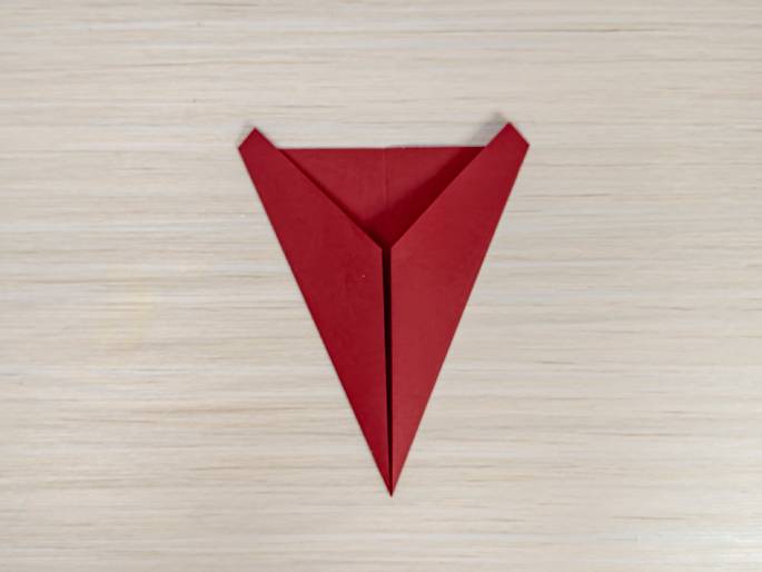 A red paper airplane, viewed from above, is centered on a light wood-grain background. The plane is simple, made from a single sheet of folded paper or cardstock. It has a pointed nose and swept-back wings. This image likely shows a stage in a paper airplane crafting process or origami.