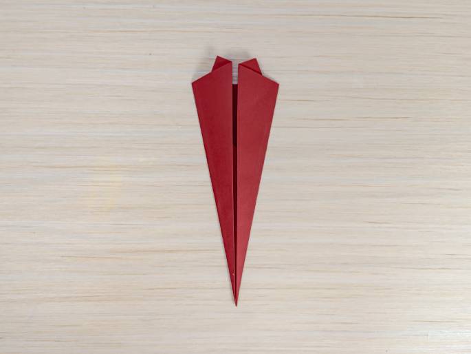 A nearly-completed red paper airplane, viewed from above, sits on a light wood-grain background. The plane's body is more elongated and narrower than in previous stages, and the wings are folded down, giving it a streamlined appearance. This image likely shows a later step in the process of making a paper airplane.