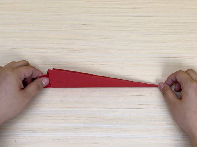 Two hands are shown manipulating a long, thin, red piece of folded paper or cardstock against a light wood-grain background. The paper resembles a flattened, elongated cone or wedge. The hands appear to be shaping or folding the paper, likely as part of a craft or origami project.