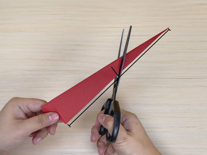 A pair of hands is using scissors to cut a long, thin, red piece of folded paper against a light wood-grained background. The paper is similar in shape to the one from the previous image, resembling a flattened, elongated cone. The scissors are cutting at an angle along the paper, likely to create a specific shape for a craft or origami project. A black line is drawn on the image, indicating the cutting line.
