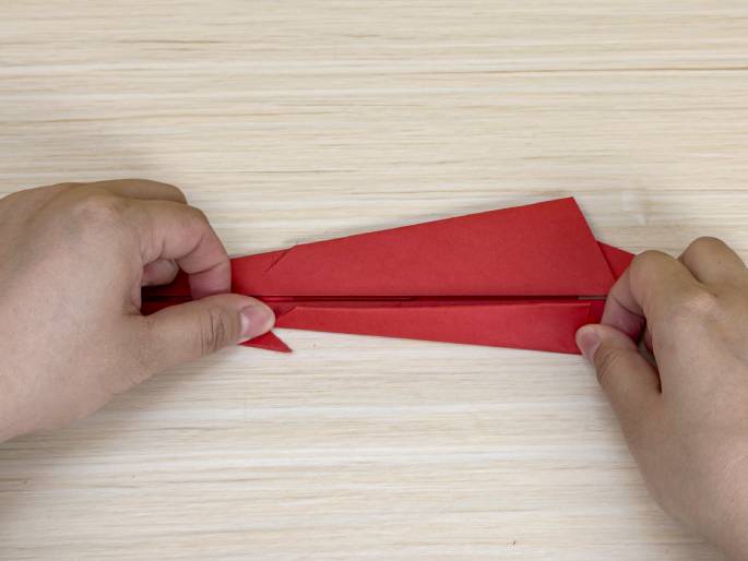 Two hands fold a red piece of paper on a light wood-grain background. The paper, which has been cut and folded into the shape of an airplane's fuselage and wings, is being held horizontally. The hands are in the process of making a small fold or adjustment near the tail section of the plane. This image likely demonstrates a step in constructing a paper airplane.