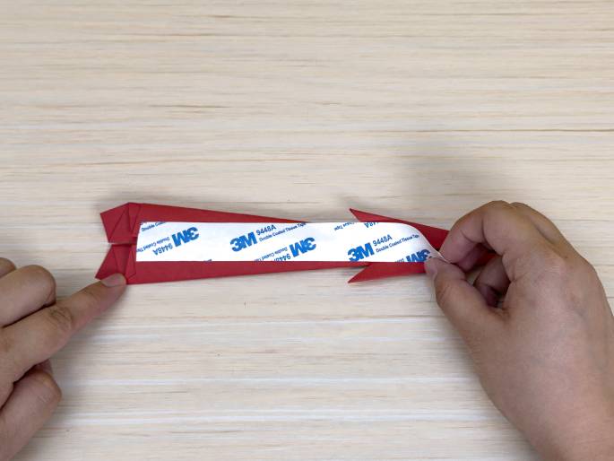 Two hands are working with a nearly-finished red paper airplane on a light wood-grain background. A piece of double-sided tape with the 3M logo is being applied to the center of the plane, likely to secure a fold or reinforce the construction. One hand holds the plane while the other peels the protective backing off the tape. This appears to be one of the final steps in the paper airplane crafting process.