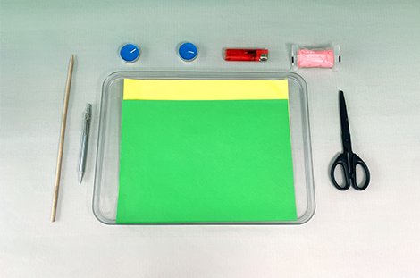 Laid out on a grey surface are the materials for a craft project: a wooden skewer, a pen, two tea lights, a lighter, pink modelling clay, green and yellow paper on a clear tray, and scissors.