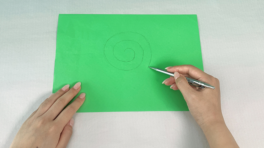 Two hands are shown, one holding a piece of bright green paper while the other draws a spiral shape on it with a pen.