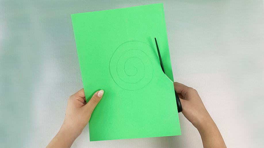 A pair of hands holds a piece of bright green paper and cuts along a pre-drawn spiral with scissors.