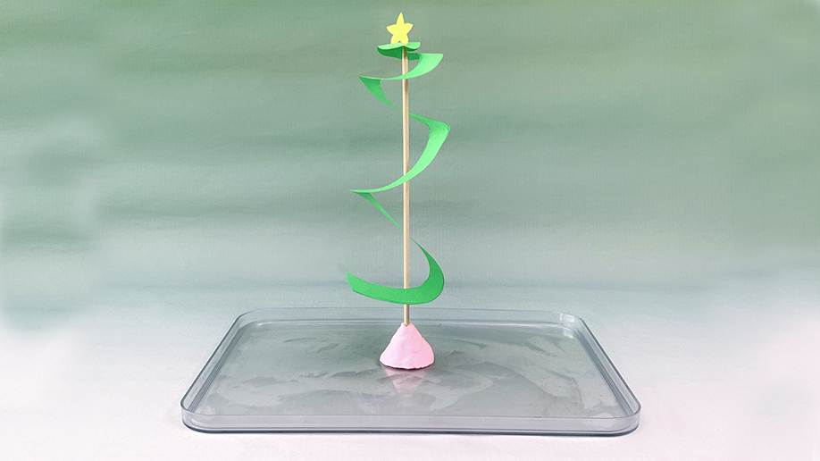 A small, spiralling Christmas tree made from green paper, topped with a yellow star and mounted on a skewer stuck into a pink clay base, sits on a clear plastic tray against a pale green background.