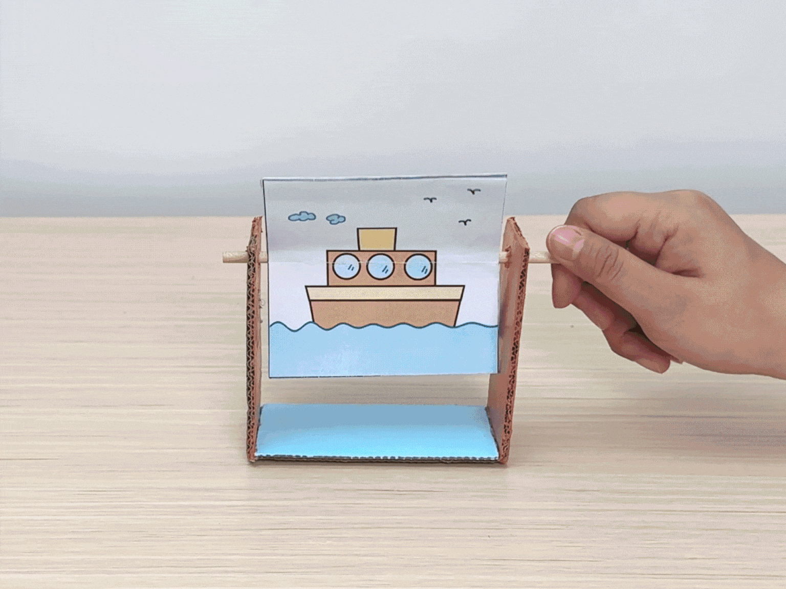 A hand turns a small, hand-cranked animation device constructed from cardboard and paper. The device has a handle made from a wooden dowel or stick. As the handle is turned, two images on separate pieces of paper rapidly alternate, creating the illusion of movement. One image depicts a boat sailing on blue water, and the other shows a white rabbit hopping across green grass. The video demonstrates a simple DIY animation toy made from readily available materials.