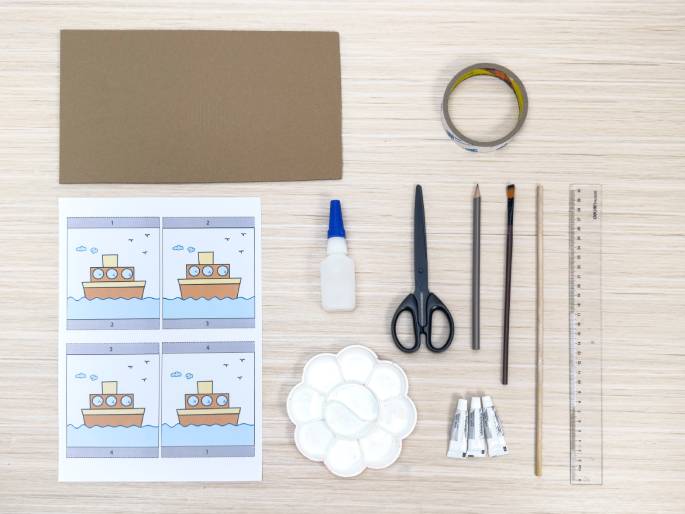 An assortment of craft supplies lies on a light wood-grained table, arranged for a children's activity. The supplies include: a sheet of brown construction paper, a roll of clear tape, a bottle of glue, scissors, a pencil, a paintbrush, a wooden dowel, a ruler, a white paint palette, and three small tubes of paint or glue. A printed sheet shows four identical cartoon images of a tugboat on water, likely a template for the craft project. The image suggests a boat-making activity for children.