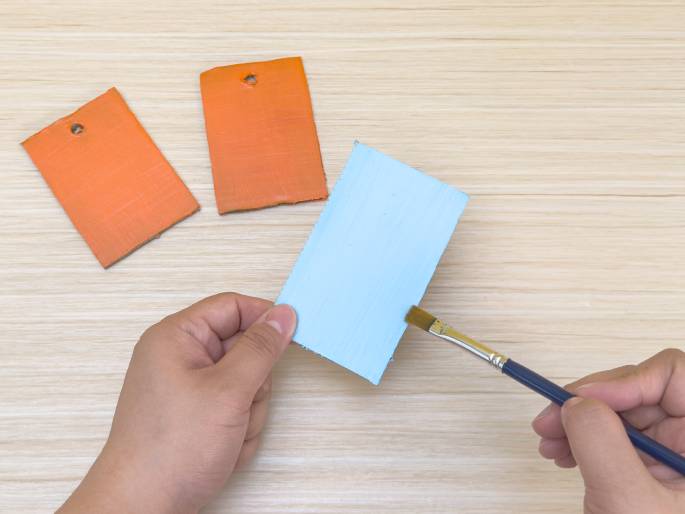 Two hands are painting a light blue rectangle of cardboard with a small paintbrush. Two other orange rectangles, similar in shape and size to the blue one, lie on the light wood-grained table nearby, suggesting they have already been painted. All three rectangles have small holes punched near their tops. This scene depicts a crafting activity, likely involving decorating these cardboard pieces.