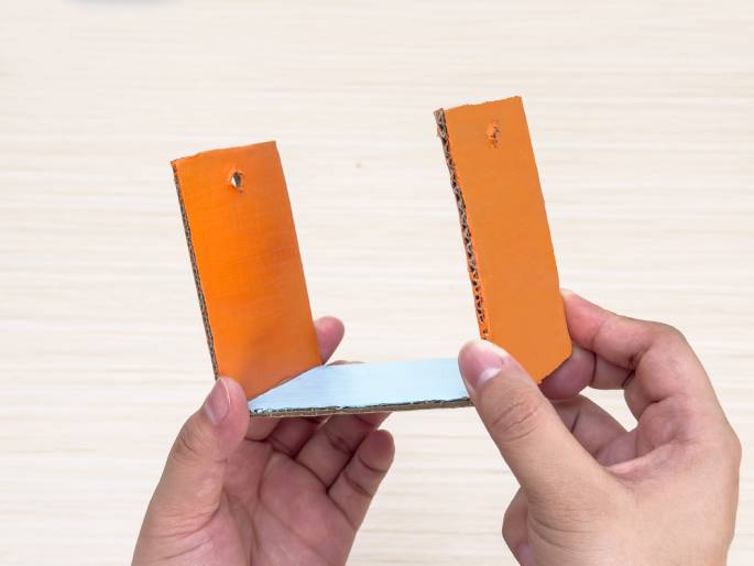 Two hands hold a partially constructed cardboard structure against a light wood-grained background. Two orange rectangular pieces, each with a hole punched near the top, are angled to form the sides of a three-dimensional shape. They are connected at the base by a light blue rectangular piece. This image shows the assembly process of a small cardboard craft.