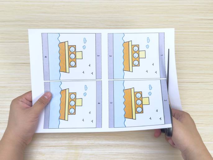 Two hands are shown working with a printed sheet of paper on a light wood-grained table. The sheet contains four identical cartoon images of a tugboat on water, arranged in a grid. One hand holds the paper while the other hand uses scissors to cut along a dotted line separating the images. This suggests the process of cutting out the individual tugboat images, likely for a craft or educational activity.