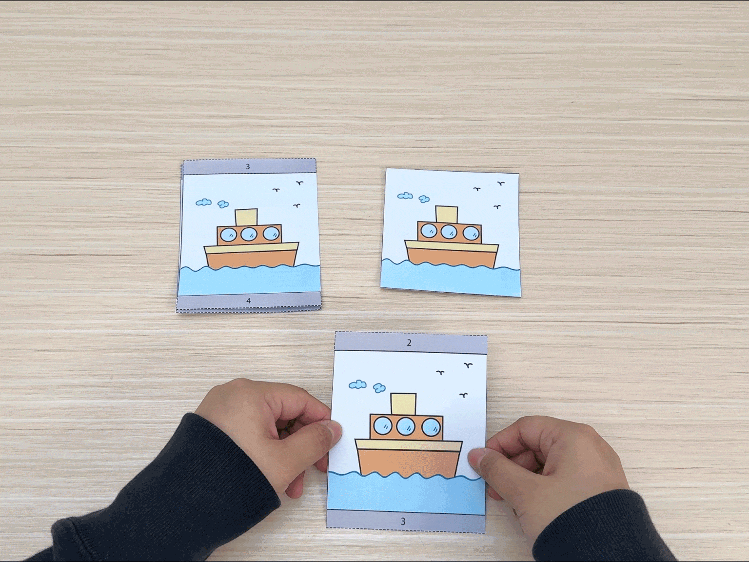 On a light wood-grained surface, two hands hold three separate square cards. Each card features a cartoon drawing of a tugboat on water, identical to the others. The hands are manipulating the cards, suggesting they've been cut from a larger sheet and are now being used for a game, sorting activity, or part of a craft project.