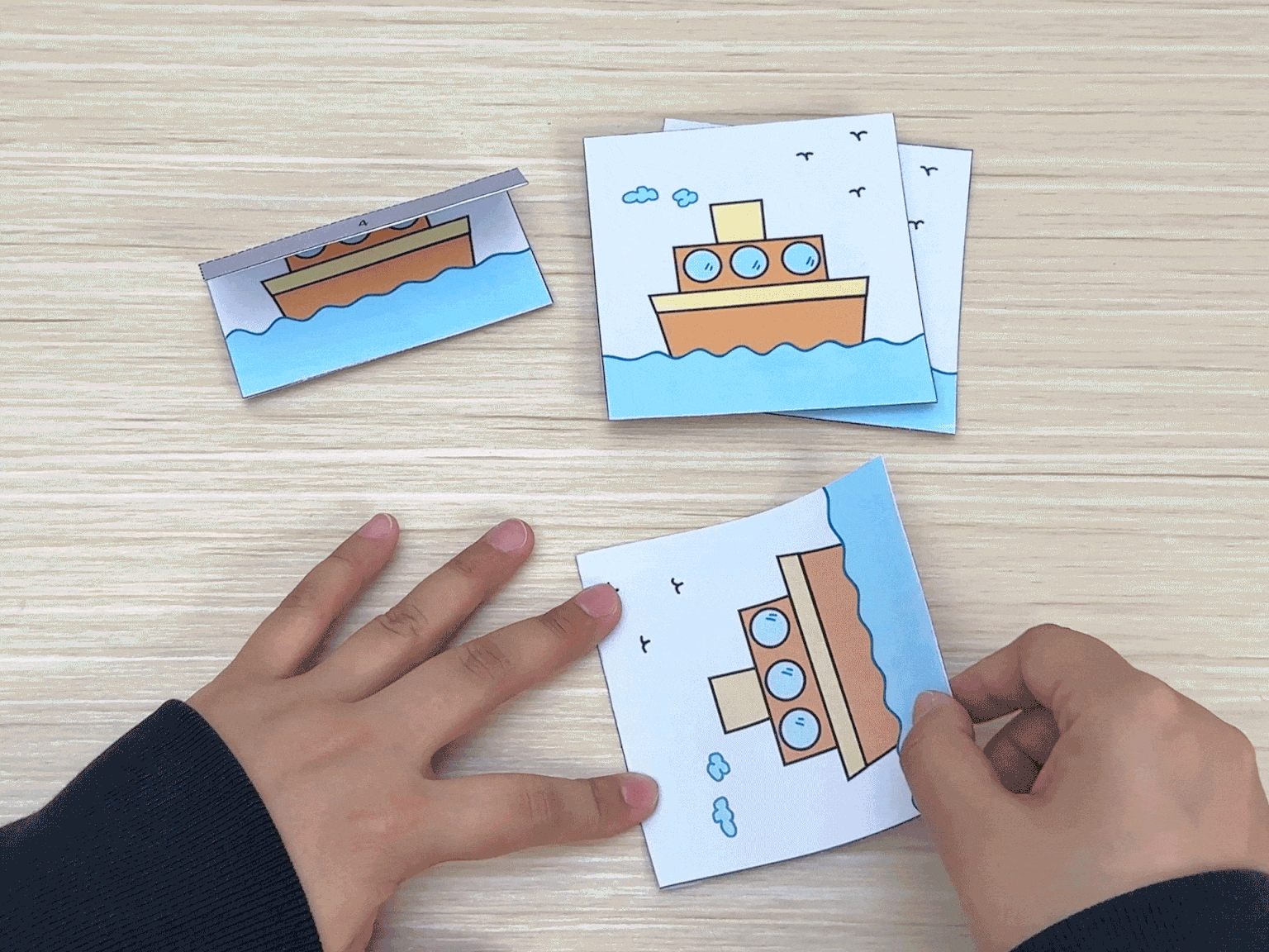 On a light wooden surface, two hands manipulate three cards featuring cartoon tugboats. One card is folded in half, another is being held flat, and the third is in the process of being folded by the hands. These actions suggest a step in a craft activity, likely involving folding and assembling the tugboat cutouts.
