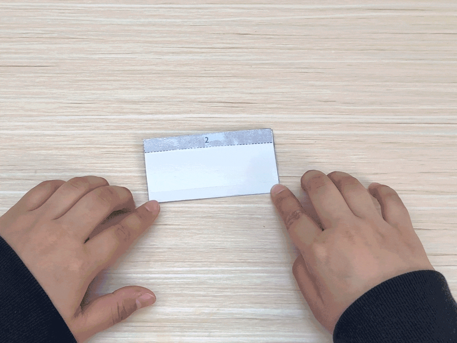Two hands are positioned on a light wood-grained table, holding a small rectangular piece of white paper. The top edge of the paper has a thin gray strip with a dotted line, indicating a fold or a section to be glued. The hands are gently pressing down on the paper, likely in the process of folding or adhering it as part of a craft project.