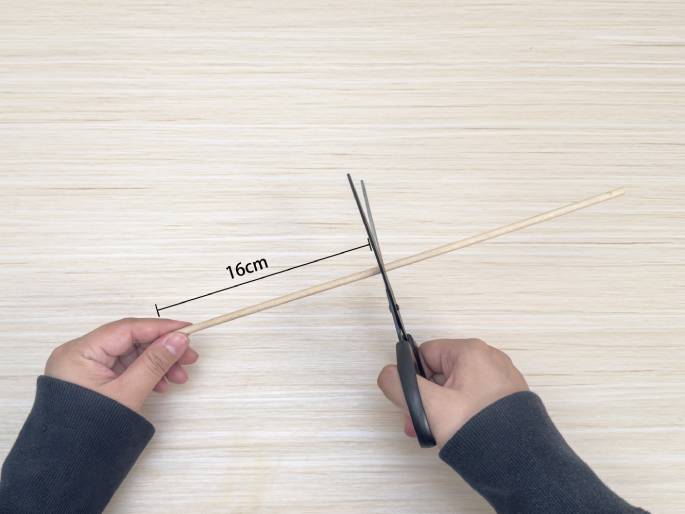 Two hands are shown cutting a thin wooden dowel with scissors on a light wood-grained table. The dowel is marked with a line indicating a length of 16cm, which is the point where the scissors are cutting. This suggests a precise measurement is being followed for a craft or construction project.