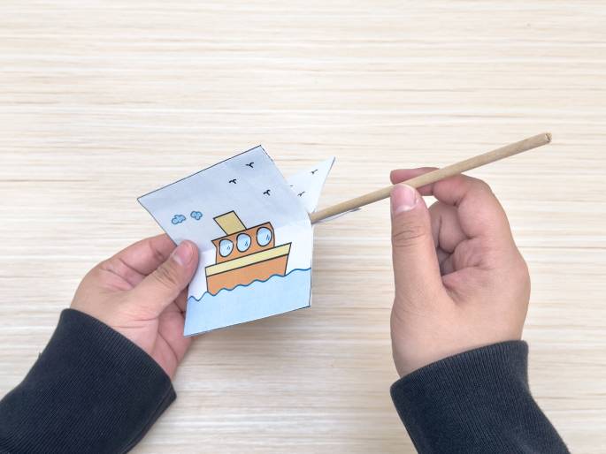 Two hands hold a folded piece of paper with a drawing of a tugboat on water and a thin wooden dowel. The dowel is inserted into the fold of the paper, creating a simple handheld boat craft. The hands are positioned above a light wood-grained table, likely the surface where this craft is being assembled.