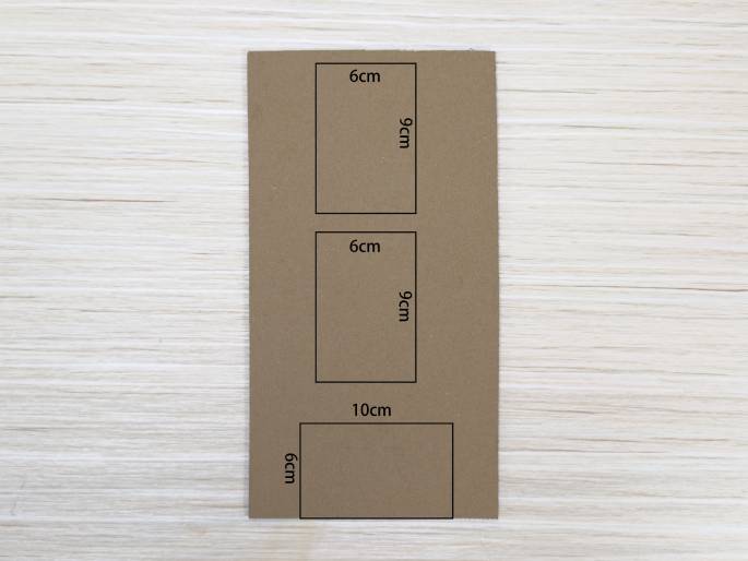 A piece of light brown cardboard lies on a light wood-grained surface. Three rectangles are outlined on the cardboard, marked with their dimensions in centimeters. Two rectangles are identical, measuring 6cm by 9cm, and the third, larger rectangle measures 6cm by 10cm. This suggests a template for cutting out shapes, likely for a craft or construction project.