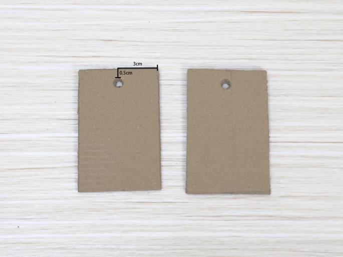Two identical light brown cardboard rectangles are placed on a light wood-grained surface. Each rectangle has a small hole punched near the top. Lines and numbers indicate the dimensions: 3cm across the top, and 0.5cm from the top edge to the center of the hole. These appear to be tags or components for a craft project.