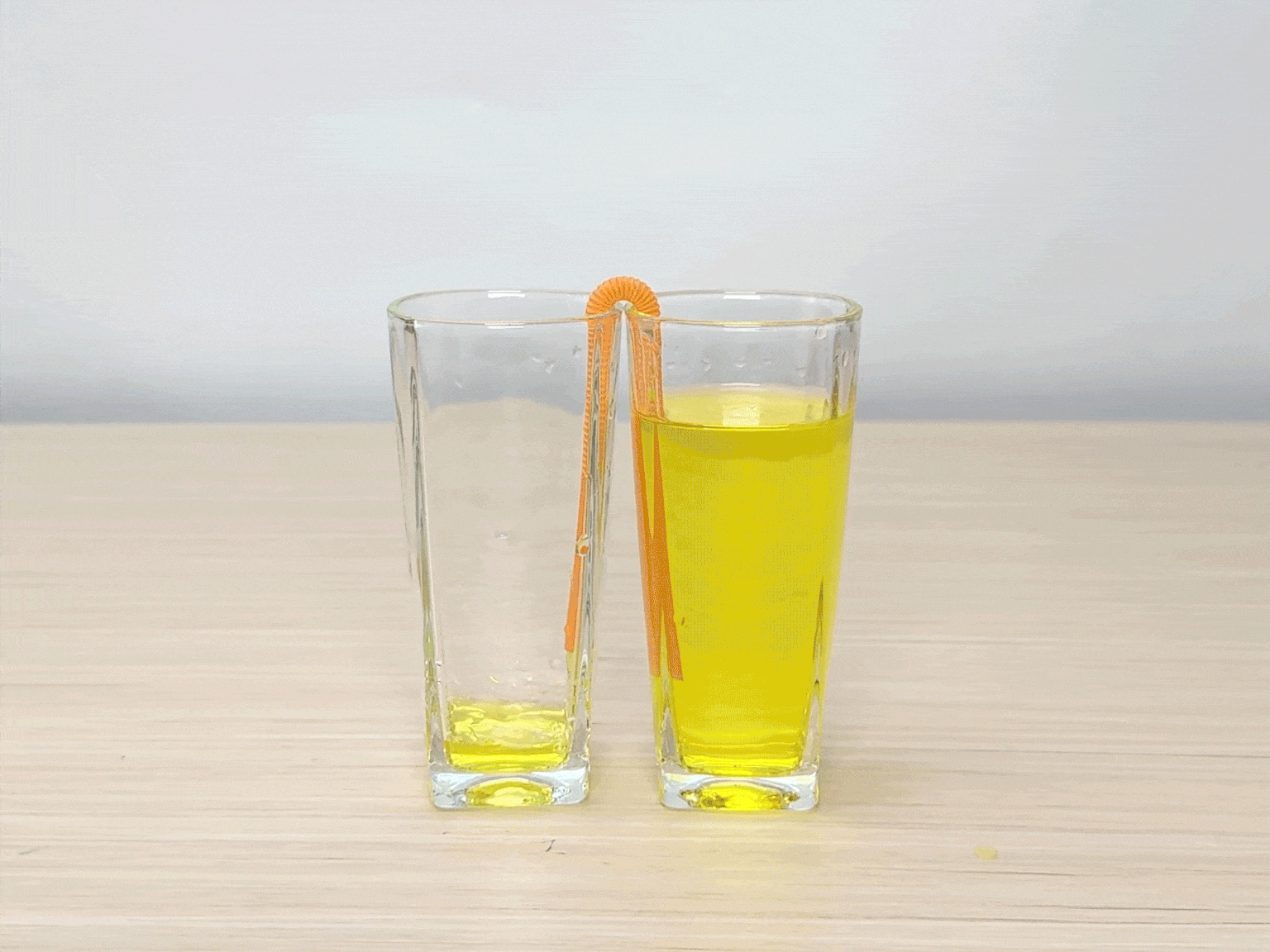Two clear glasses, partially filled with a bright yellow liquid, sit side-by-side on a wooden table. A flexible orange drinking straw is placed so that one end is submerged in the liquid of each glass. The video appears to be demonstrating a simple science experiment, possibly illustrating the principles of siphoning or capillary action.
