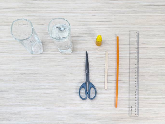 An overhead flatlay image displays various craft supplies arranged on a light wood grain surface. The items include two clear glasses (one empty, one with water), a small bottle of yellow liquid, a pair of scissors, a wooden craft stick, an orange plastic drinking straw, and a clear plastic ruler. This image depicts the materials needed for a craft project.