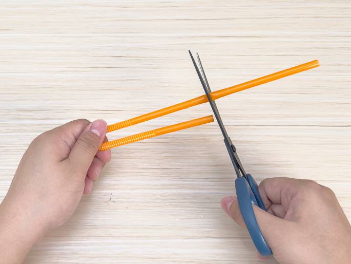Two hands are shown cutting an orange plastic drinking straw with a pair of scissors. One hand holds the straw against a light wood grain surface while the other hand operates the scissors. The straw has a textured, ridged section near where it is being cut. This image shows the process of cutting a straw for a craft project.