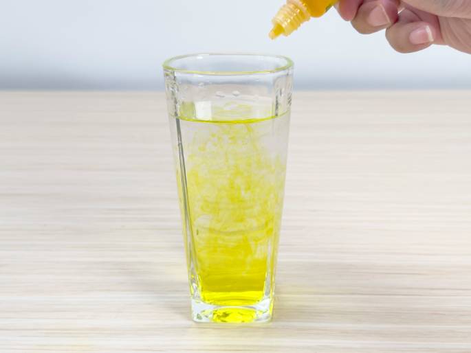 A hand squeezes a small, clear bottle containing yellow liquid into a glass of water. The yellow liquid is dispersing into the water, creating a cloudy, bright yellow effect. The glass sits on a light wood grain table against a plain off-white background. This image likely depicts food coloring or another dye being added to water.