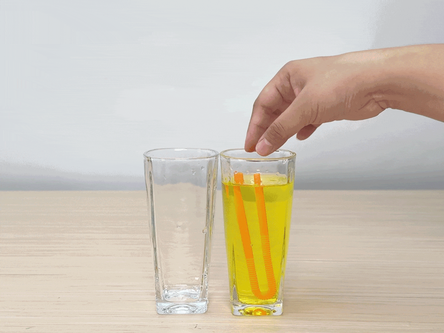 A hand places two segments of an orange drinking straw into a glass of bright yellow liquid. An empty clear glass sits beside it on a light wood grain surface. The background is a solid off-white. This image appears to be a step in a craft or science experiment.