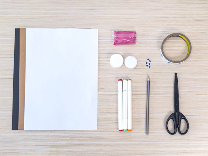 Laid out on a light wood-grain surface are various art supplies, likely for a children's craft project. On the left is a stack of rectangular paper: a white sheet on top of brown and dark gray sheets. To the right of the paper are, from top to bottom: a small, sealed package of pink modeling clay, two white bottle caps, a small pile of googly eyes, a roll of clear tape, three white markers with red, green, and orange caps, a gray pencil, and a pair of black scissors. The arrangement suggests preparation for a crafting activity.